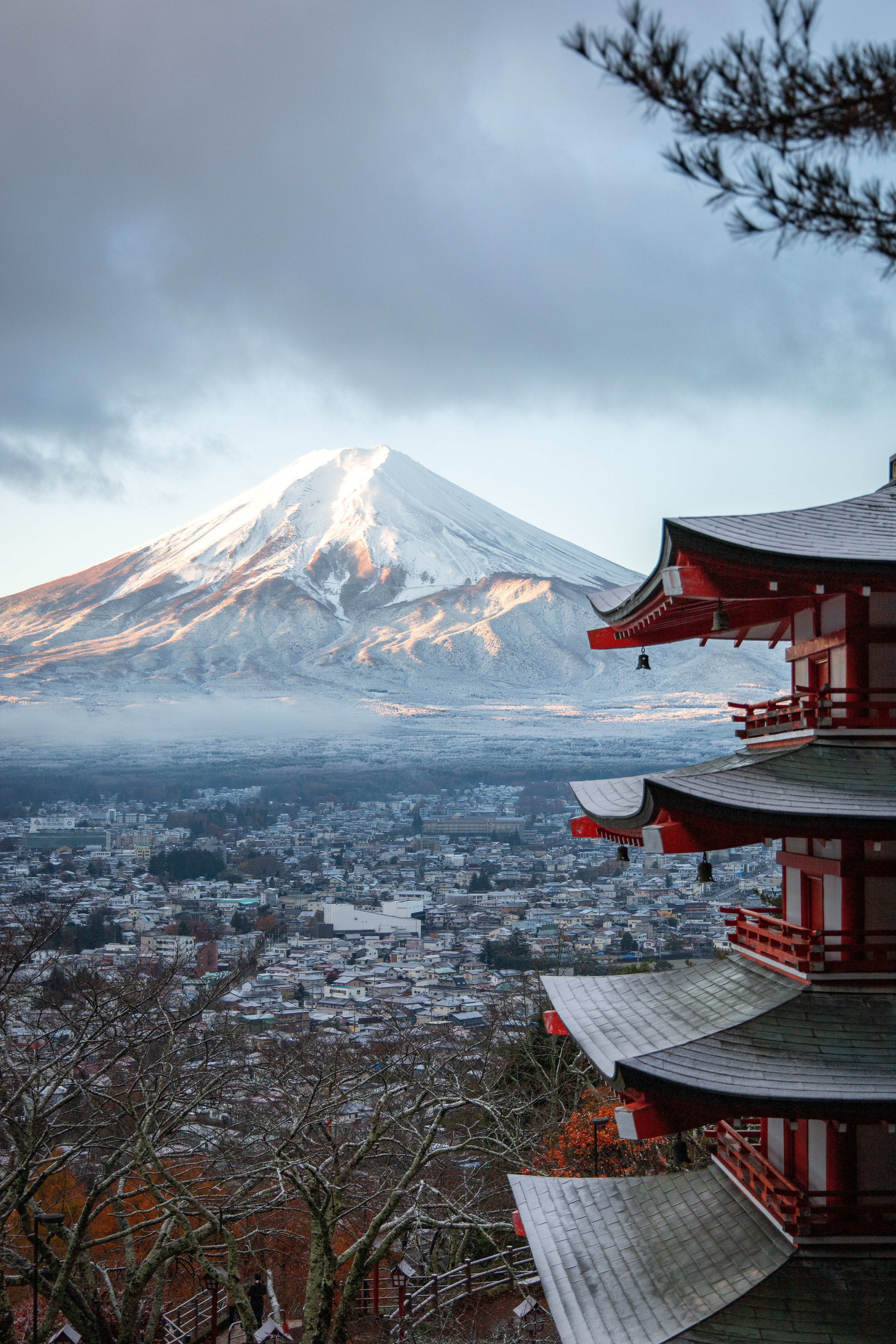 Zdjęcie przedstawia górę Fuji i miasto położone poniżej. Kadr jest zrobiony z daleka. Na pierwszym planie, po prawej stronie widoczny jest czerwony, drewniany budynek. Jest widoczna jedynie jego część, a dokładniej 3 boczne dachy ułożone jeden nad drugim. Nad najwyższym dachem można zauważyć gałęzie drzewa z uschniętymi liśćmi. Góra Fuji znajduje się w centrum zdjęcia, w głębi. Jest bardzo dobrze widoczna. Szczyt góry jest przyprószony śniegiem. Poniżej widać skały oświetlone 1 promieniem słońca, a poniżej wstążkę mgły. Niebo nad górą jest jasno niebieskie ale z lewej strony widać nadciągające ciemno granatowe chmury. U podnóża gór widac rozległe miasto. W panoramie miasta, nie odznacza się żaden budynek. Wszystkie widoczne zabudowania są kwadratowe niskie i pokryte białym kolorem. Być może to śnieg. Cały krajobraz przywodzi na myśl początek zimy z chłodem i małą jeszcze ilością śniegu.