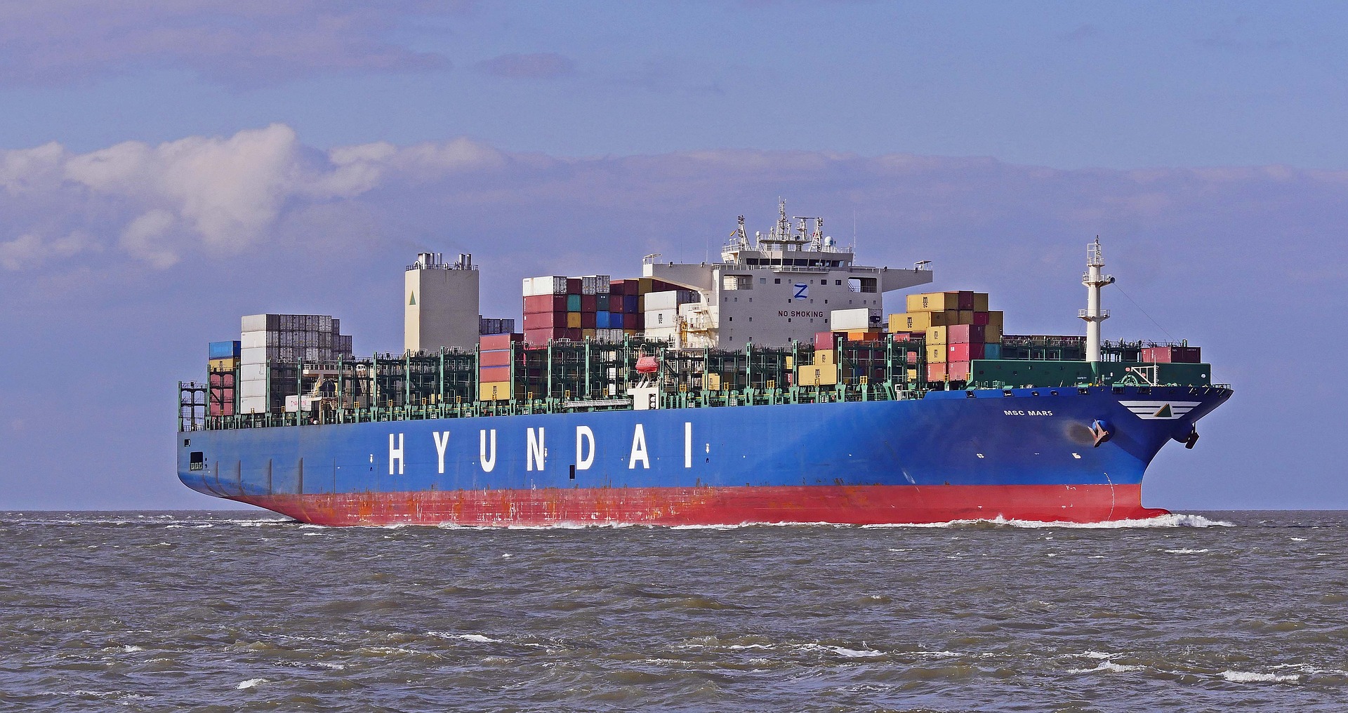 Na zdjęciu statek -kontenerowiec. Na prawej burcie - powyżej linii zanurzenia ma napis Hyundai. KOntenerowiec jest na morzu. Morze w kolorze jest szare, niebo niebieskie z chmurami. Na statku widać poukładane różnokolorowe kontenery oraz mostek kapitański. Burty statku są pomalowane na granatowo, napis jest biały, a część zanurzająca się w wodzie jest czerwona
