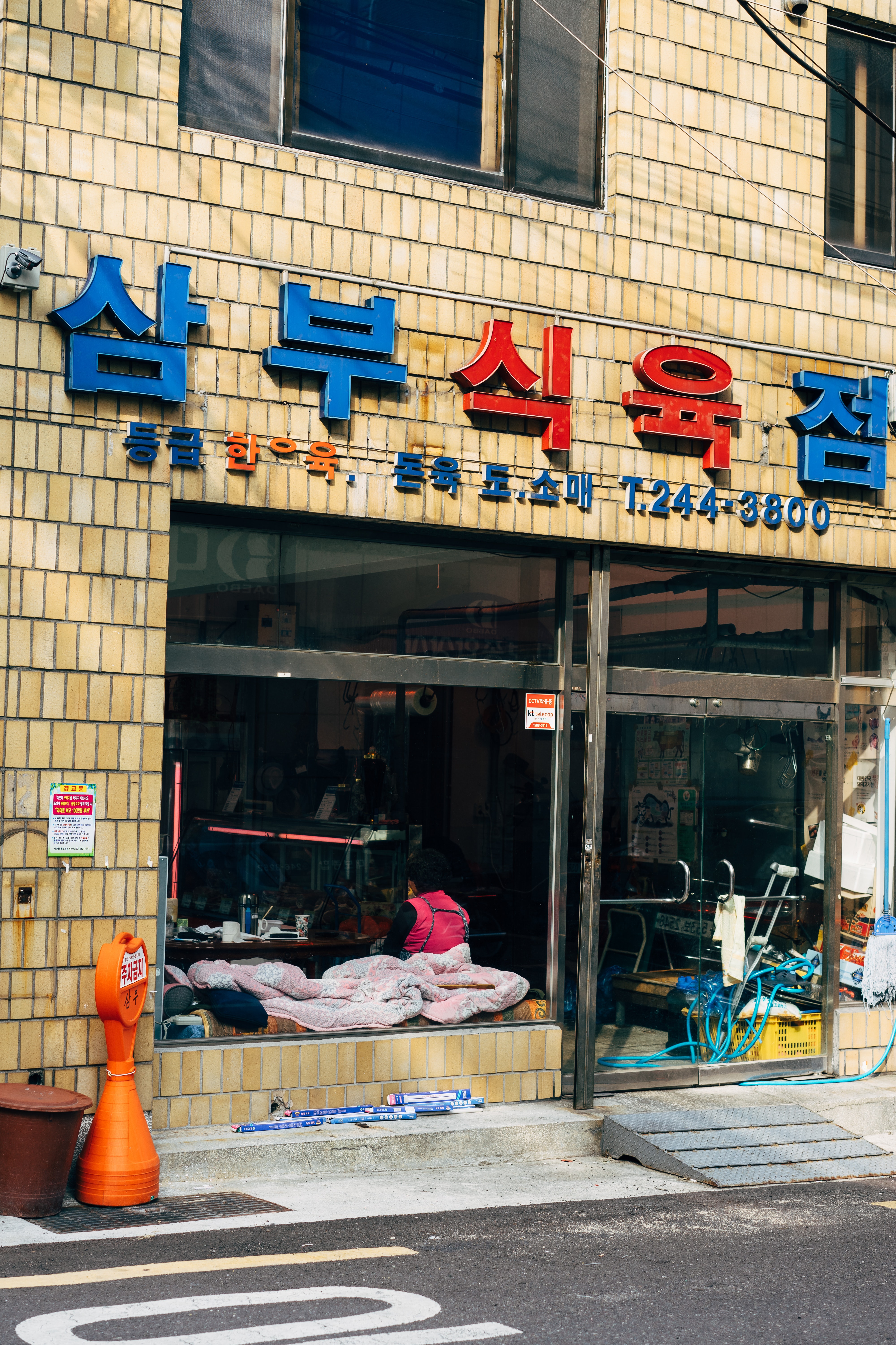 Zdjęcie przedstawia ścianę domu mieszkalnego (ściana z cegiełek w kolorze piasku pustyni), w którym na parterze jest bar lub restauracja. Nad wejściem wisi 5 znaków w języku koreańskim. Znaki środkowe są czerwone, pozostałe niebieskie. Witryna sklepowa i drzwi wejściowe są duże i przestronne.