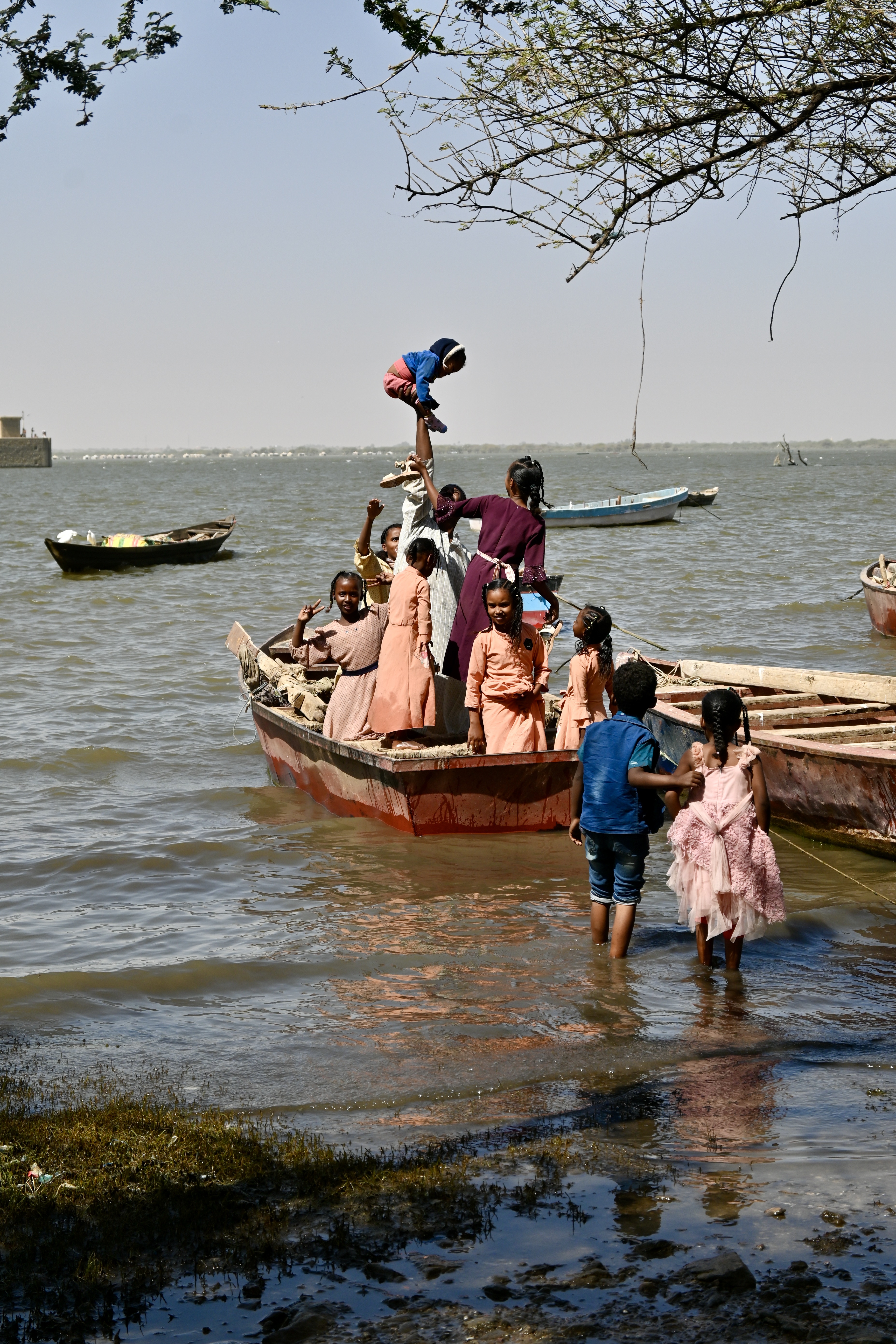 Miejsce, gdzie zostało zrobione zdjęcie: Dżebel Aulia nad Nilem Białym w Sudanie. Autorem zdjęcia jest Maciej Kurcz. Na zdjęciu widać wodę, która może być morzem, jeziorem - a w rzeczywistości jest to rzeka Nil Biały. Na zdjęciu nie widać drugiego brzegu, woda sięga po widnokrąg. Woda jest szaro-niebieska, z elementami żółtymi przy brzegu. Jest spokojna przy brzegu można zobaczyć lekki zarys fal. Na wodzie stoi płaskodenna łódka, w niej zasiada rodzina, która prawdopodobnie wyrusza na wycieczkę. W łódce siedzi już m.in 6 osób a podchodzi jeszcze 2 dzieci. Wszystkie osoby, są ubrane w kolorowe stroje. Kobiety maja sukienki w odcieniach różu, beżu i kolorze herbacianym. Jedno z dzieci ma granatową bluzę, a jedna osoba bordową. Obok łódki, która wyrusza na wycieczkę stoi druga pusta. W oddali na wodzie widać jeszcze 2 inne zacumowane puste łodzie. Niebo jest niebieskie, a całe zdjęcie robi wrażenia bardzo optymistyczne i wesołe.