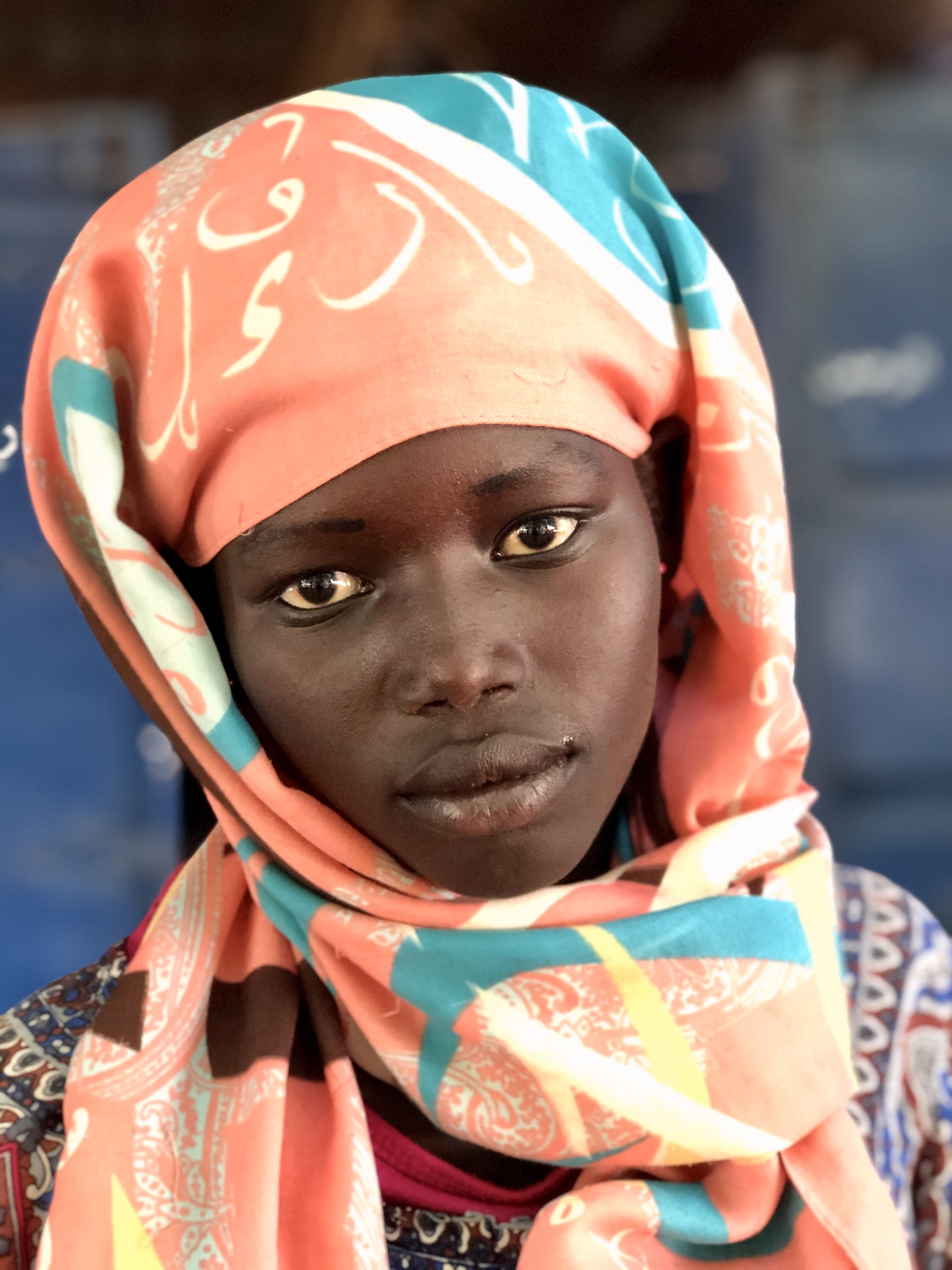 Zdjęcie zostało wykonane w miejscowości Soba w Sudanie. Autorem zdjęcia jest pan dr hab, Maciej Kurcz, prof. UJ. Na zdjęciu ukazana jest głowa kobiety - dziewczynki o czarnym kolorze skóry. Twarz jest młoda, lekko uśmiechnięta, jakby zamyślona. Dziewczyna ma spokojne oczy ciemnobrązowe, drobny nos i stosunkowo pełne usta również jak cala twarz brązowe. Na głowie dziewczyna ma kolorowy turban, który obejmuje głowę i spada luźno na barki wiązanie. Turban jest zasadniczo w kolorach łososiowo- błękitnym ze wzorem z liter alfabetu arabskiego. Tło zdjęcia jest ciemno-niebiesko szare..