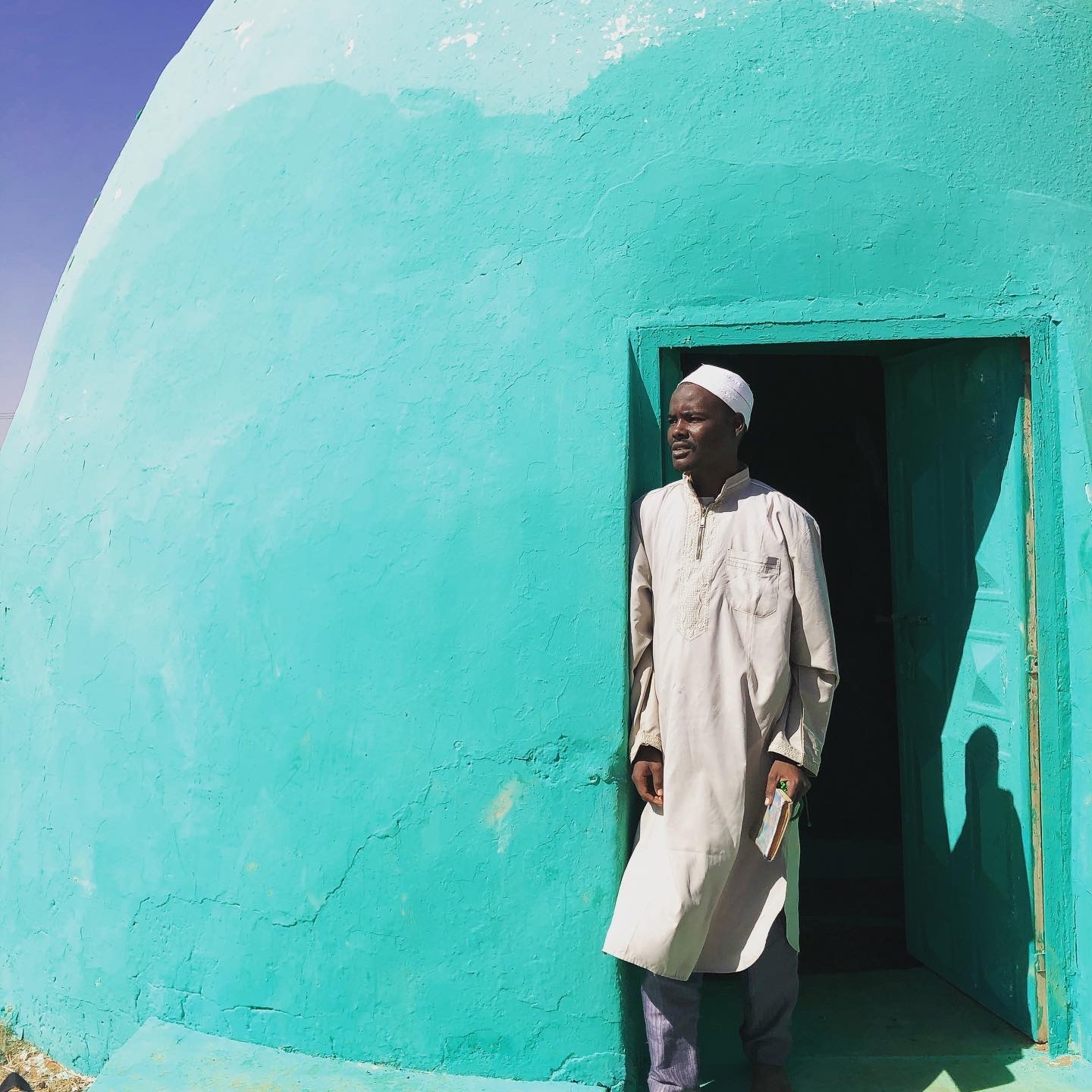 Zdjęcie wykonane jest w miejscowości Soba w Sudanie Południowym. Autor zdjęć: Maciej Kurcz Zdjęcie jest piękne kolorystycznie. Dominują 3 kolory biały, lazurowy i błękitny. Na zdjęciu widać po lewej stronie u góry skrawek błękitnego nieba. Większą część zdjęcia wypełnia kopulasty grobowiec - grób świątobliwego mędrca. Budowla ma kształt owalu po prawej stronie znajdują się prostokątne drzwi do świątyni, w których stoi mężczyzna. Mężczyzna jest derwiszem opiekującym się tą światynią, który za opłtą odmawia Koran za wiernych. Mężczyzna ma czarny kolor skóry jest ubrany w białą, długą, prostą szatę zapinaną pod szyję na 3 guziczki. Na głowie ma białą czapeczkę - kufi.